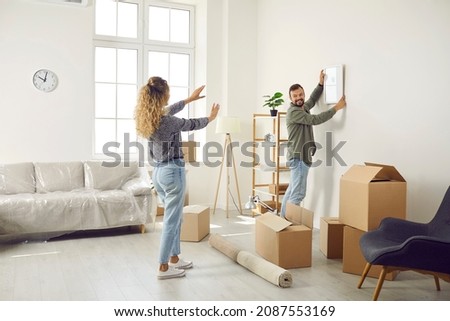 Happy married couple who recently bought house are unpacking stuff and decorating new home. Young man and woman are making living room cozy and choosing place on wall to hang painting or family photo 商業照片 © 