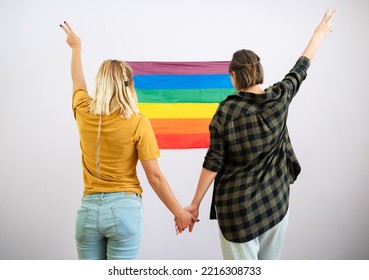 Happy Married Homosexual Female Gay Couple Holding Hands With Rainbow Pride Flag On Background - Lesbian Couple At Home Decoreting Living Room