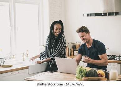 Happy married interracial couple looking at laptop computer together while doing daily moring routines in the kitchen at home