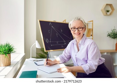 Happy Math Teacher In The School Classroom. Portrait Of A Beautiful Elegant Middle Aged Woman In A Shirt And Glasses Sitting At Her Working Desk, Taking Some Notes, Looking At The Camera And Smiling