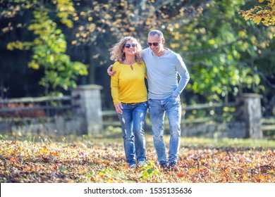 Happy Mature 40s Couple In Autumn Park In The Embrace Walk On The Fallen Maple Leaves.