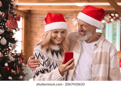 Happy mature 50 years old couple wear santa hats using phone near Christmas tree at home buy presents online ecommerce shopping in mobile app, have New Year virtual party video call meeting on cell. - Powered by Shutterstock