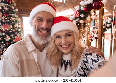 Happy Mature 50 Years Old Grandparents Couple In Santa Hats Waving Hands Greeting Family On Distance Video Call, Having Remote Online Virtual Xmas Party Meeting On Christmas At Home. Webcam View