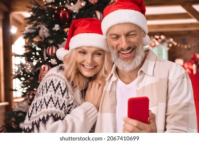 Happy mature 50s family couple wear santa hats using cell phone stand near Christmas tree at home do holiday online e commerce shopping in app, have xmas virtual party video call meeting on cellphone. - Powered by Shutterstock