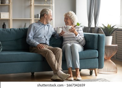 Happy Mature 50s Husband And Wife Sit Rest On Comfortable Sofa In Living Room Enjoy Tea Talking, Smiling Elderly 60s Couple Relax On Couch At Home Drink Coffee Chat Speak Laugh On Leisure Weekend