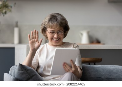 Happy mature 60s woman waving hand hello at tablet screen, speaking on video call, talking to family online, smiling, laughing, sitting on sofa at home. Distant communication concept - Powered by Shutterstock