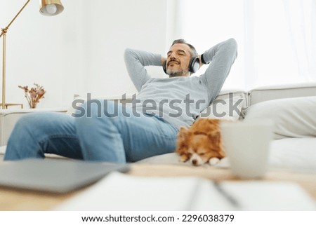 Happy Mature Adult man sitting on sofa relaxing  and listening to music with headphones in living room at home. Senior male sleeping on couch with his chihuahua dog. Well-being, lifestyle, recreation