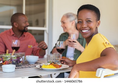 Happy Mature African Woman Looking At Camera While Having Lunch With Her Friends In Background. Portrait Of Laughing Black Woman Enjoying Brunch With Smiling Senior Couple Eating And Drinking Together