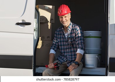 Happy Mature Artisan Sitting In Van With Cardboard Box And Paint Buckets. Smiling Mature Man With Hardhat And Work Gloves Ready For Work. Portrait Of Painter Sitting With Box In Van For Moving House.