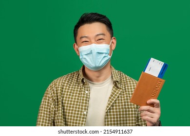 Happy Mature Asian Man In Medical Mask Holding Plane Tickets And International Passport, Posing On Green Studio Background. Male Traveling Overseas During Coronavirus Lockdown