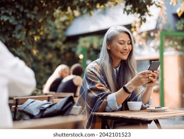 Happy mature Asian woman surfs internet on mobile phone sitting at small table on outdoors cafe terrace on autumn day - Powered by Shutterstock