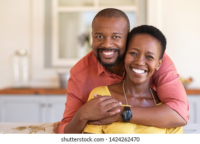 Happy mature black couple bonding to each other and smiling while sitting on couch. Portrait of smiling black man embrace his wife from behind and looking at camera.