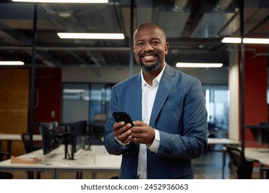 Happy mature black man in businesswear looking at camera while using mobile phone in office - Powered by Shutterstock