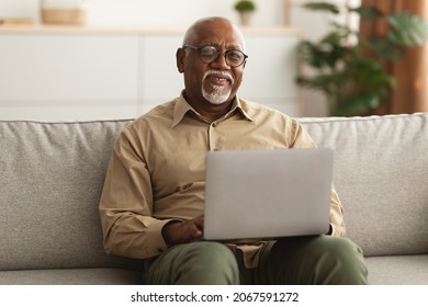 Happy Mature Black Man Using Laptop Browsing Internet Smiling To Camera Working Online Sitting On Sofa At Home, Wearing Glasses. Freelance In Older Age, E-Learning For Senior People