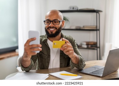 Happy Mature Black Man Using Credit Card And Smartphone While Shopping Online From Home, Sitting At Desk With Laptop. Man Making Payment Via Bank App On Phone. E-commerce Concept