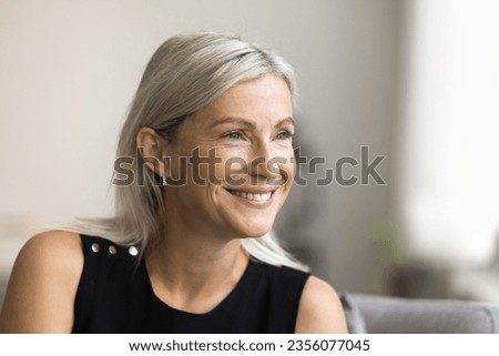 Happy mature blonde lady looking away with toothy smile, thinking, dreaming, posing at home, laughing in good thoughts. Positive senior woman close up candid facial portrait