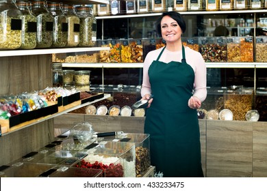 Happy Mature Brunette Woman Wearing Apron And Selling Nuts And Dried Fruits In Organic Shop
