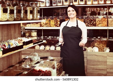 Happy Mature Brunette Woman Wearing Apron And Selling Nuts And Dried Fruits In Organic Shop
