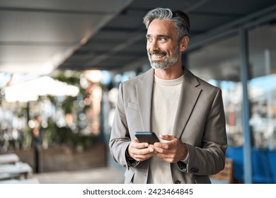 Happy mature business man executive using mobile cell phone corporate tech outdoors. Smiling handsome middle aged professional businessman holding cellphone looking away standing outside. Copy space