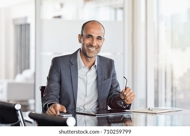 Happy mature business man holding spectacles in modern office. Successful senior businessman sitting in meeting room with phone and tablet. Smiling man in suit at elegant office.