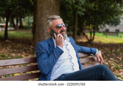 A Happy Mature Business Man Talking On Mobile Phone While Siting At The Park Bench.