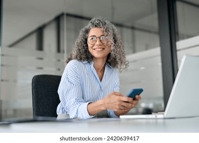 Happy mature business woman executive holding mobile cell phone looking away in office. Smiling middle aged professional businesswoman manager wearing glasses using cellphone working on smartphone. - Powered by Shutterstock