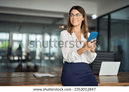 Happy mature business woman executive holding cell phone looking away in office. Smiling mid aged 40s professional businesswoman manager entrepreneur using cellphone working on smartphone. Copy space