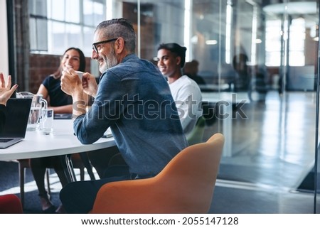 Happy mature businessman attending a meeting with his colleagues in an office. Experienced businessman smiling cheerfully while sitting with his team in meeting room. Businesspeople working together