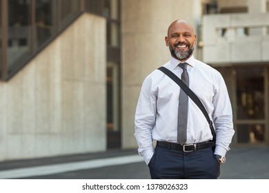 Happy Mature Businessman With Laptop Bag Standing On Street. Smiling Business Man In Formal Clothing Looking At Camera In The City Centre. Black Man Going To Work With Building In The Background.