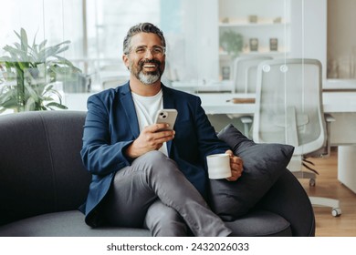 Happy, mature businessman sitting on a grey sofa, holding a cup of coffee and texting on his smartphone. Senior entrepreneur dressed in business casual in a professional office. - Powered by Shutterstock