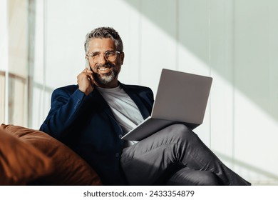 Happy, mature businessman talking on his phone while holding his laptop, is comfortably seated in a modern office. Senior entrepreneur engaging in business communications. - Powered by Shutterstock