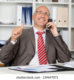 Happy mature businessman using landline phone while gesturing thumbs up at office desk
