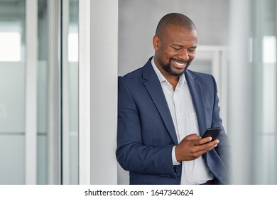 Happy Mature Businessman Using Smartphone In Modern Office. Smiling African American Business Man Reading Message On Cellphone. Middle Aged Man In Formal Using Mobile Phone In Workplace.