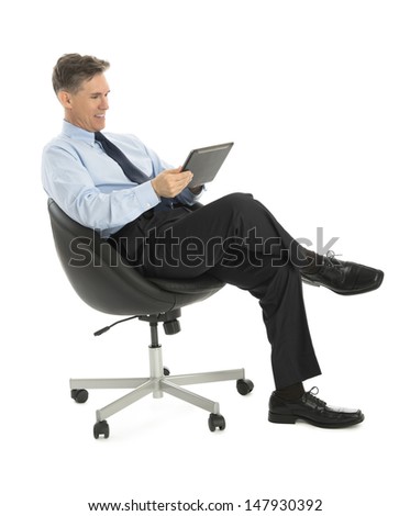 Happy mature businessman using tablet computer while sitting on office chair against white background