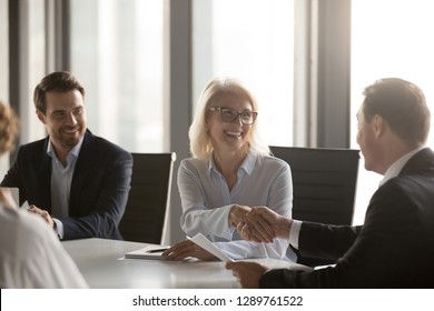 Happy Mature Businesswoman Handshaking New Male Partner, Ceo And Smiling Middle Aged Lady Shaking Hands At Group Meeting Making Deal, Rewarding Or Hiring, Celebrating Business Contract With Client