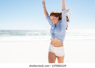 Happy Mature Casual Woman Enjoying Tropical Beach Vacation. Smiling Young Woman Having Fun On Her Vacation At Sea. Joyful Lady With Red Hair Enjoying Freedom With Outstreched Arms, Jumping On Beach.