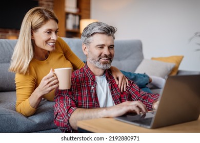 Happy Mature Caucasian Wife And Husband Looking At Laptop, Enjoy Coffee Break In Free Time In Living Room Interior. Video Call, Meeting Online, Modern Technology, Social Network And Blog At Home