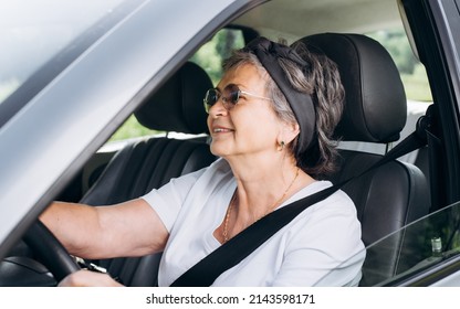 Happy Mature Caucasian Woman Driving Car While Sitting At Wheel Inside Interior Of Vehicle. Smiling, Cheerful Senior Woman Driver Wearing Seat Belt. Active Elderly People.