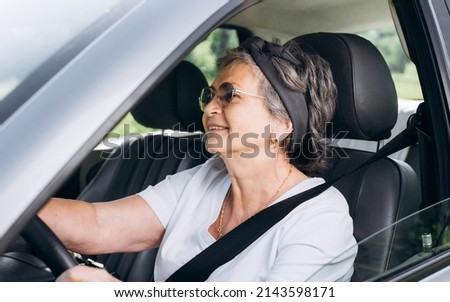 Happy mature caucasian woman driving car while sitting at wheel inside interior of vehicle. Smiling, cheerful senior woman driver wearing seat belt. Active elderly people.