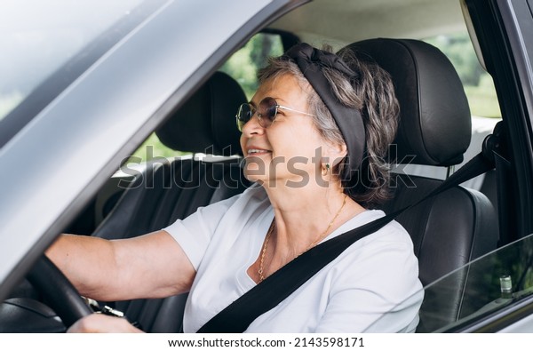 Happy mature caucasian
woman driving car while sitting at wheel inside interior of
vehicle. Smiling, cheerful senior woman driver wearing seat belt.
Active elderly people.