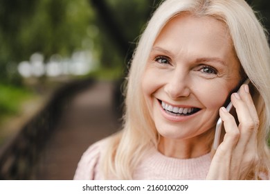 Happy Mature Caucasian Woman Talking On Cellphone, Calling To Her Friends, Ordering Food Delivery, Taxi On Smart Phone, Looking At Camera In Park Forest.