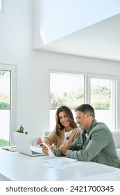 Happy mature couple calculating money expenses savings using laptop at home. Middle aged man and woman checking receipts for tax refund, paying bills online sitting at table in living room. Vertical.