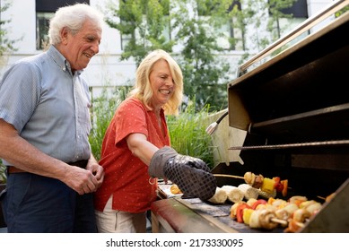 Happy Mature Couple Cooking Kebabs Outside On Their Grill.