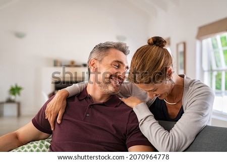 Happy mature couple embracing on sofa while laughing. Middle aged woman hugging husband from behind while smiling and joke. Beautiful wife and mid adult man having fun and enjoying time together.