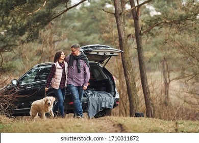 Happy mature couple have a walk with their dog in autumn or spring forest near modern car.