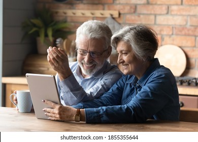 Happy mature couple having fun with tablet, sitting in kitchen, laughing senior man wearing glasses drinking tea or coffee in morning, grandparents chatting with relatives online, browsing apps - Powered by Shutterstock