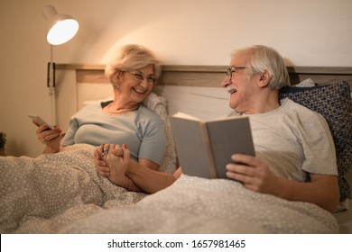 Happy Mature Couple Holding Hands While Resting In Bed And Communicating In The Evening. 