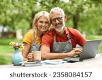 Happy Mature Couple With Laptop And Tea Relaxing In Garden, Senior Spouses In Aprons Using Computer And Having Coffee While Sitting At Terrace Of Their Country House, Enjoying Retirement Leisure