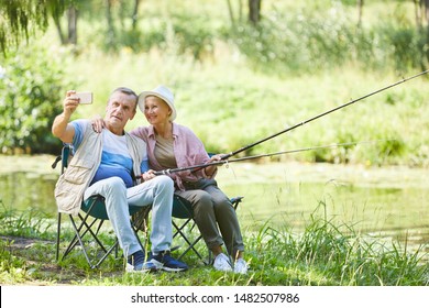 Happy Mature Couple Making A Selfie Portrait On Mobile Phone While Fishing Together Near The River In Nature