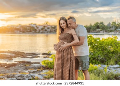 A happy, mature couple over 40, enjoying a leisurely walk on the waterfront On the Sunset, their joy evident as they embrace the journey of pregnancy later in life - Shutterstock ID 2372844931